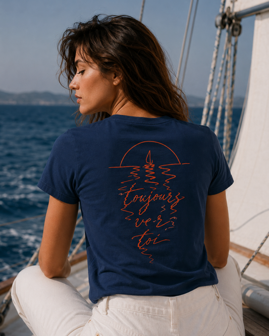 Back view of the Je Reviendrai navy tee worn on a sailing boat. Coral illustration of a sailboat at sunset with the words toujours vers toi. L'Ame Sirène collection by muzee.