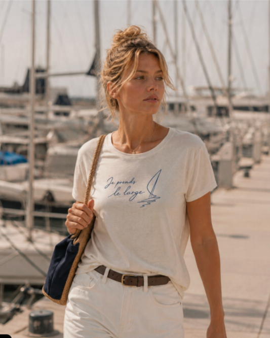 Cream fitted t-shirt with navy handwritten 
"Je prends le large" script and sailboat 
illustration, worn by a woman at a sunny 
Mediterranean marina with yacht masts in 
the background