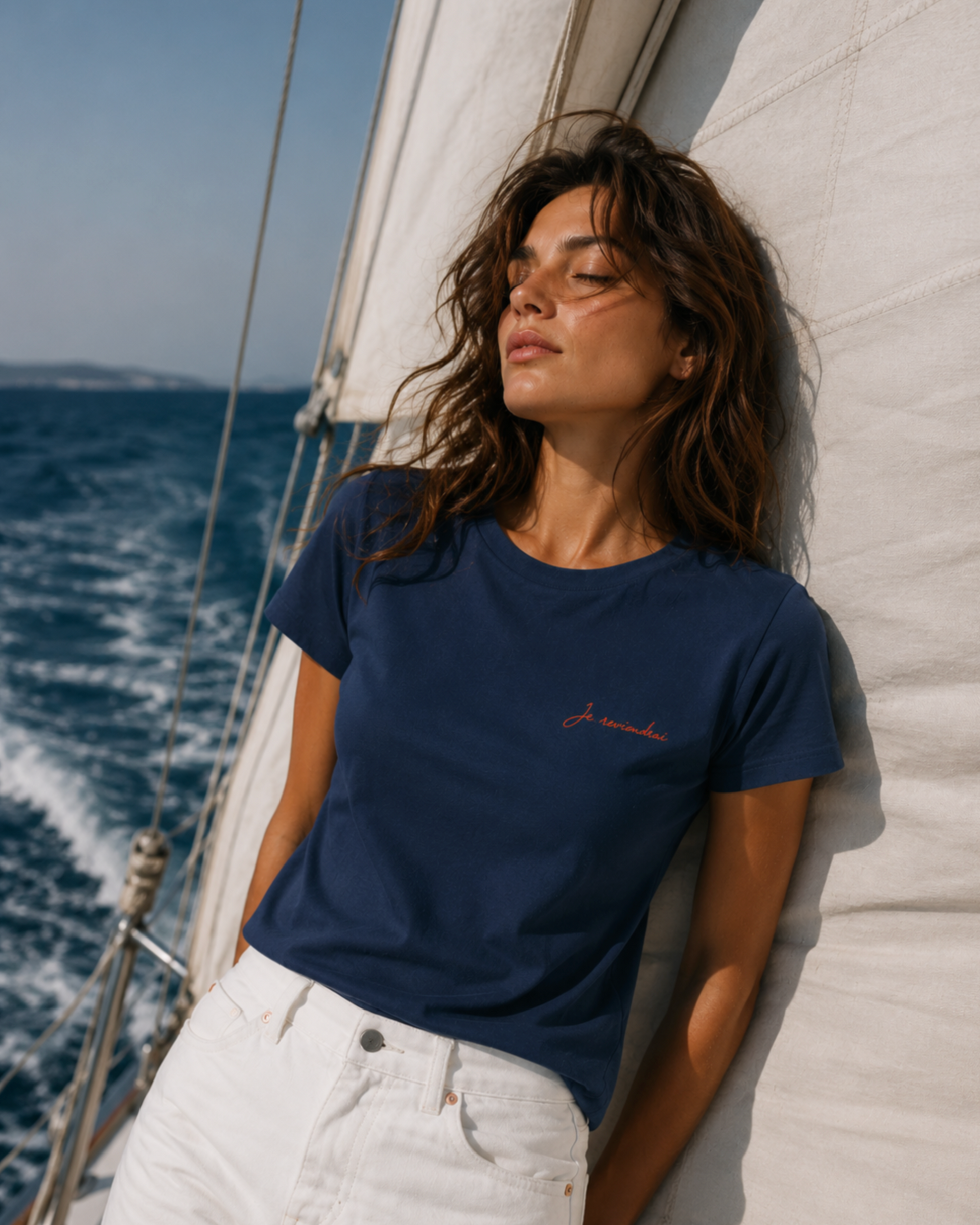 Woman wearing the Je Reviendrai navy tee on a sailing boat. Coral cursive print on the left chest. L'Ame Sirène collection by Muzee.