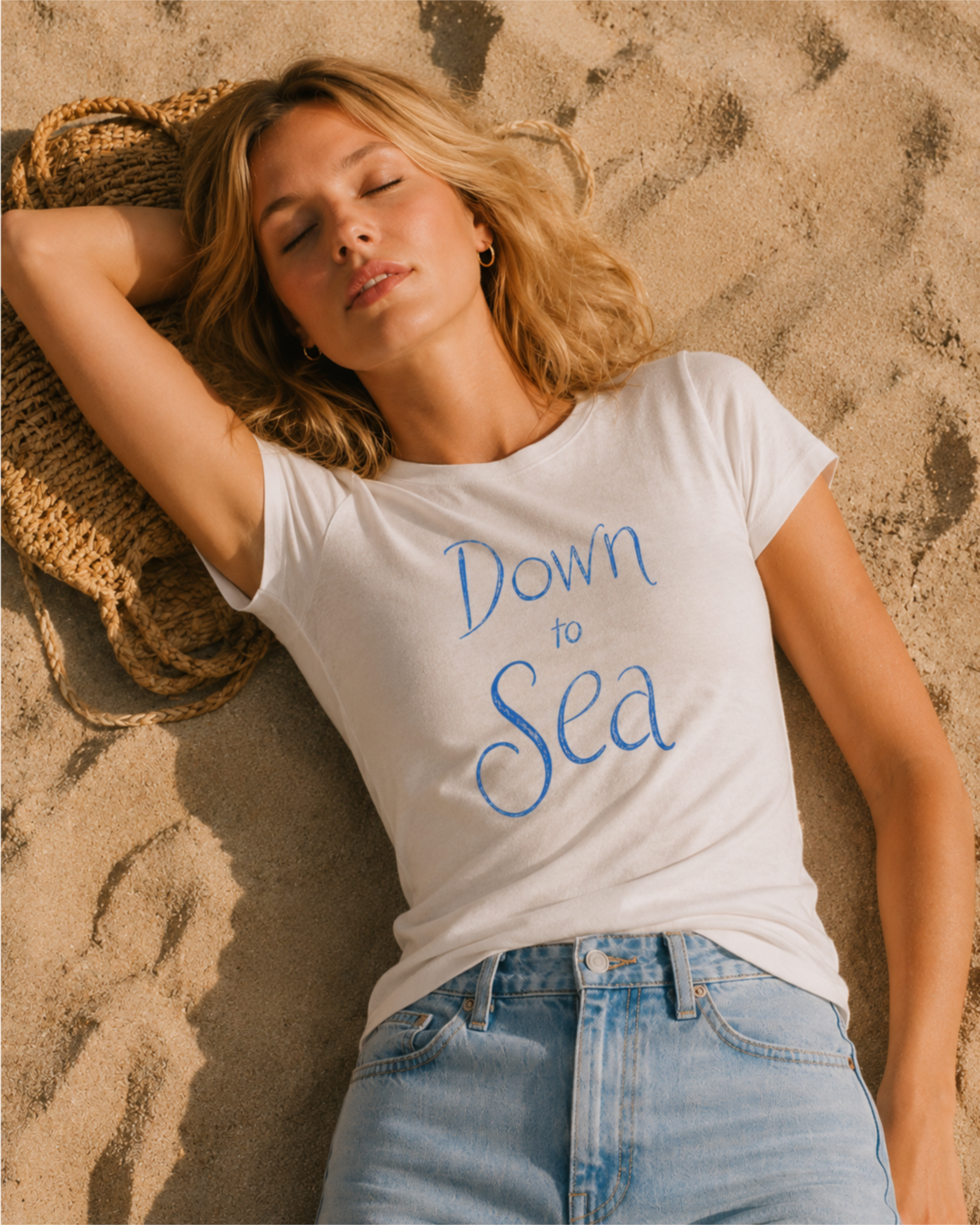 Editorial beach photo of a woman lying on sand in a white summer T-shirt with a blue “Down to Sea” print.
